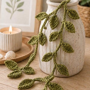 May include: A decorative arrangement featuring a cream-colored ceramic vase with two strands of green crocheted leaves cascading down. A lit candle and framed botanical print are in the background, creating a natural, handcrafted aesthetic.