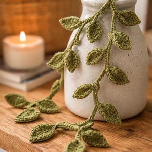 May include: A handmade, green crochet leaf garland draped over a white ceramic vase. The leaves are detailed and textured, with a natural, organic aesthetic. The vase sits on a wooden surface, with a lit candle and a woven basket in the background.