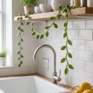 May include: A long, green crocheted vine garland featuring small, textured leaf shapes. The yarn plant decoration is draped across a wooden kitchen shelf, with two long strands hanging down, showcasing intricate stitch details in a vibrant green.