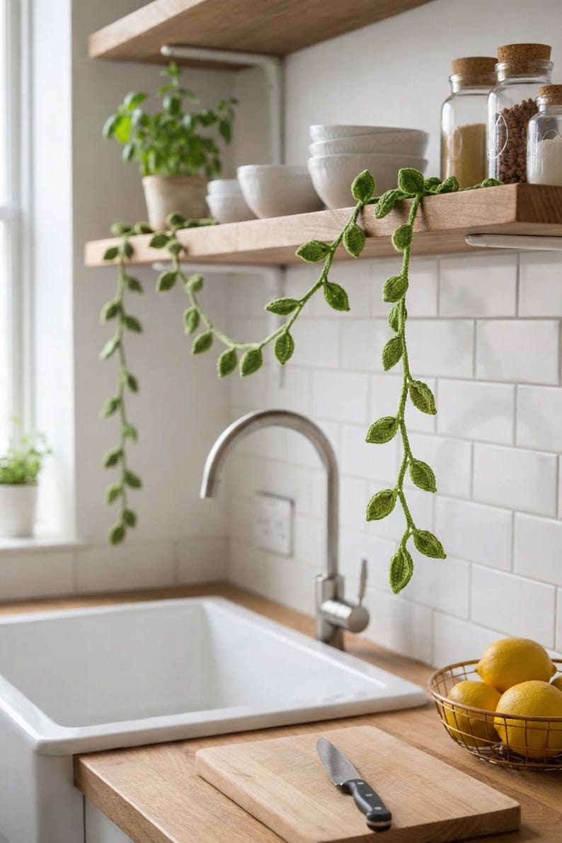 May include: A long, green crocheted vine garland featuring small, textured leaf shapes. The yarn plant decoration is draped across a wooden kitchen shelf, with two long strands hanging down, showcasing intricate stitch details in a vibrant green.