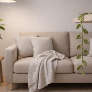 May include: A crocheted vine with small green leaves hanging down from a wooden shelf. The vine features a series of individual leaf shapes connected in a vertical strand, crafted with a textured yarn finish in a light olive green color.