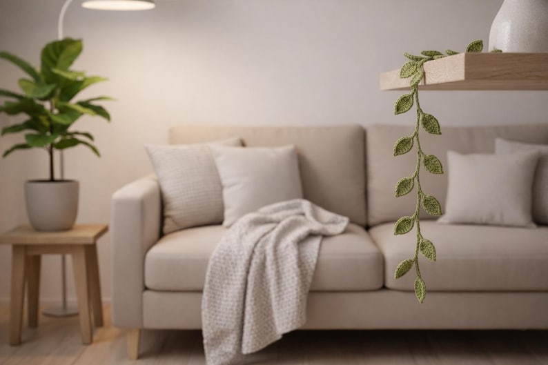 May include: A crocheted vine with small green leaves hanging down from a wooden shelf. The vine features a series of individual leaf shapes connected in a vertical strand, crafted with a textured yarn finish in a light olive green color.