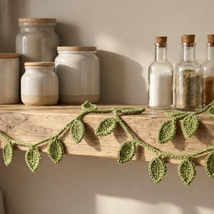 May include: A green crocheted garland featuring a series of small, textured leaves connected by a thin yarn vine. The garland is draped horizontally across the front edge of a wooden shelf, positioned in front of several ceramic jars and glass bottles.