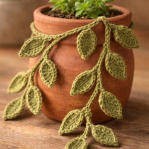 May include: A terracotta clay pot containing a small green succulent plant, adorned with a decorative crocheted vine featuring multiple light green leaves that drape over the sides of the round, earthy-toned container.