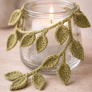 May include: A clear glass candle holder wrapped with a crocheted vine featuring sage green leaves. The vine is draped around the top rim of the cylindrical glass vessel, which contains a single white candle with a lit flame in the center.