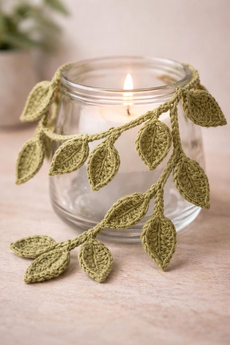 May include: A clear glass candle holder wrapped with a crocheted vine featuring sage green leaves. The vine is draped around the top rim of the cylindrical glass vessel, which contains a single white candle with a lit flame in the center.