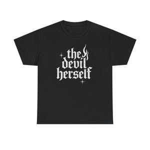 Puede incluir: Camiseta negra con el texto gótico blanco "the devil herself". El diseño incluye llamas y estrellas. La camiseta es de cuello redondo.