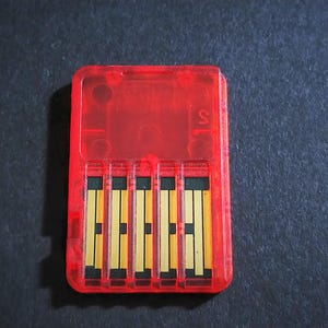 Könnte beinhalten: Eine transparente rote Spielkassette mit gold- und schwarzfarbenen Anschlüssen. Die Kassette hat eine rechteckige Form mit abgerundeten Ecken und ist vor einem dunklen Hintergrund platziert. Der Buchstabe "S" ist auf der Kassette sichtbar.