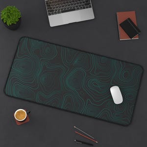 May include: A black desk mat with a teal topographic design. A white computer mouse sits on the mat. A laptop, a small potted plant, a cup of coffee, a notebook, and writing utensils are also on the dark surface.