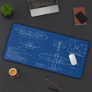May include: A blue desk mat with a detailed aircraft blueprint design. The mat features technical drawings, measurements, and text. A white computer mouse sits on the mat, with a laptop, notebook, and a coffee cup nearby.