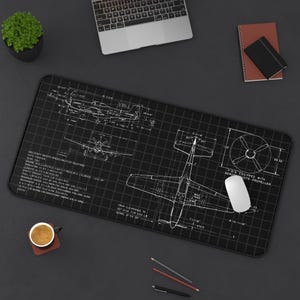 May include: Black rectangular desk mat with a white blueprint of a P-51 Mustang aircraft. The design includes technical drawings, measurements, and text. A white computer mouse rests on the mat, alongside a cup of coffee and a laptop.