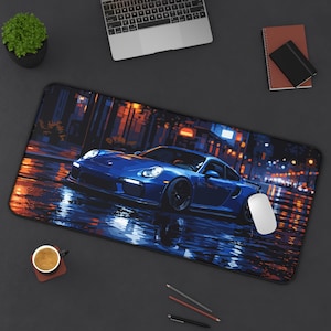 May include: A rectangular desk mat featuring a blue sports car on a wet street at night. The car is the focal point, with reflections in the water. A white mouse is on the right side of the mat. A laptop, plant, and coffee cup are also visible.