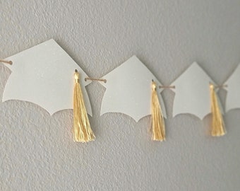 May include: A white graduation cap banner with gold tassels. The banner is strung together with twine and is perfect for a graduation party.