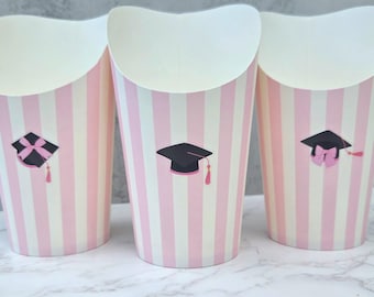 Pode incluir: Três copos de papel listrados em rosa e branco para guloseimas. Cada copo apresenta um gráfico de chapéu de formatura com uma borla rosa. Os copos têm uma borda superior curva e são adequados para guardar lanches ou pequenos presentes.
