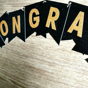 Pode incluir: Faixa preta e dourada com a palavra "CONGRATS" escrita em letras douradas com glitter.
