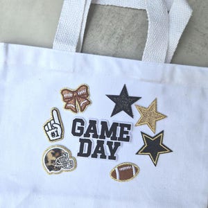 May include: A white canvas tote bag featuring the text GAME DAY in black block letters. The bag is decorated with embroidered patches including a brown football, a leopard print helmet, a foam finger, a bow, and three glittery stars.