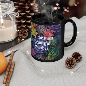 Puede incluir: Taza de cerámica negra con un diseño de suculentas coloridas y el texto "For the most Beautiful Mother". La taza tiene un asa curva y contiene una bebida humeante. Elementos decorativos rodean la taza.