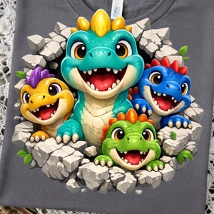 May include: A dark grey t-shirt featuring a graphic print of four colourful cartoon dinosaurs emerging from a cracked stone wall. The dinosaurs are teal, yellow, blue, and green, each with distinct spikes and wide, happy, open-mouthed expressions.