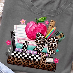 May include: A dark grey t-shirt featuring a graphic of a stack of books, a bright pink apple, a pencil, and a ruler. The design includes a large leopard print bow, a chequered pattern on the book spines, and small pink flower accents.