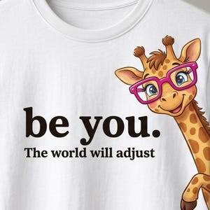 May include: A white t-shirt featuring a cartoon giraffe wearing bright pink glasses. The giraffe is positioned on the right side of the shirt. To the left, the text reads: be you. The world will adjust. The design is printed in black and pink.