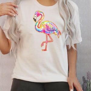 May include: White t-shirt featuring a colorful flamingo design. The flamingo has a pink body, a rainbow-colored wing, and a gold glitter outline. The bird is standing on one leg.