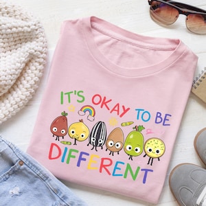 Può includere: T-shirt rosa chiaro con la scritta colorata "IT'S OKAY TO BE DIFFERENT". La maglietta presenta una grafica di frutta e verdura a fumetti sorridenti. La maglietta è realizzata in un materiale morbido.