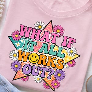 May include: A light pink t-shirt featuring a large, multi-colored star graphic with the text "WHAT IF IT ALL WORKS OUT?" in bold, bubble-style lettering. The design includes a yellow smiley face, various colorful daisies, and patterns.