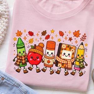 May include: A light pink t-shirt featuring a graphic of five anthropomorphic school supplies including a green crayon, a red apple wearing a knit hat, an orange glue stick, a floral notebook, and a yellow pencil, all wearing scarves and boots.