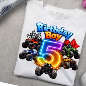 May include: A white t-shirt featuring a graphic print with the text Birthday Boy and a large glowing number 5. The design includes four colorful monster trucks, checkered flags, and red flags arranged around the central number 5 graphic.