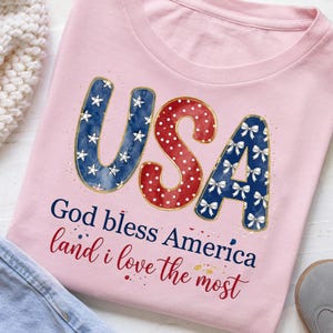 Op de afbeelding: Een lichtroze t-shirt met de tekst USA in grote letters met een blauw sterrenpatroon, rode stippen en een blauw strikpatroon. Daaronder staat de tekst God bless America and land i love the most in donkerblauwe en rode letters.