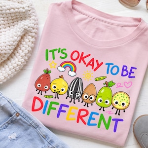 Puede incluir: Una camiseta rosa claro con un gráfico colorido y el texto IT'S OKAY TO BE DIFFERENT. El diseño incluye ilustraciones de dibujos animados de varias semillas y frutas con caras, un pequeño arcoíris, un sol, crayones y un símbolo de corazón.