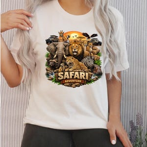 May include: White t-shirt featuring a colorful Safari Adventure design. The graphic includes a lion, elephant, giraffe, zebra, cheetah, and other animals, with the words "Safari Adventure" in a wooden-style font. The design is set against a sunset backdrop.