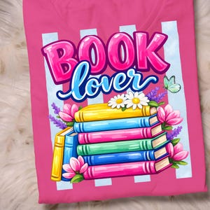 May include: A bright pink t-shirt featuring a graphic of a stack of colorful books with the text "BOOK lover" in bold pink and blue script. The design includes white daisies, purple lavender sprigs, pink flowers, and a small green butterfly.
