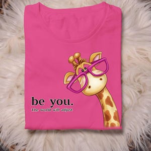Op de afbeelding: Een felroze t-shirt met een cartoon giraffe met een roze bril. De giraffe kijkt opzij. De tekst "be you. The world will adjust" staat eronder in het zwart gedrukt.
