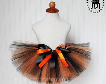 Puede incluir: Un tutú de tul negro y naranja con un lazo de satén negro. El tutú está en un maniquí blanco.