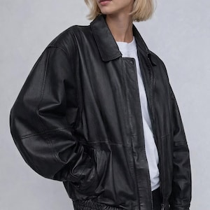 Peut inclure: Veste de bombardier en cuir noir avec un col, des poches avant et un ourlet côtelé. La veste a une fermeture éclair et est portée sur un t-shirt blanc.