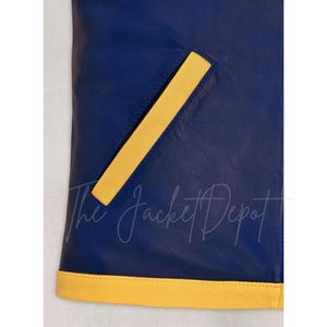 May include: Close-up of a section of a blue leather jacket with a yellow pocket and trim. The jacket has a smooth texture and a classic design. The text "The Jacket Depot" is visible.