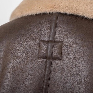 Puede incluir: Primer plano de una chaqueta de cuero marr&oacute;n con cuello de piel sint&eacute;tica beige. La chaqueta tiene una superficie texturizada y un detalle de parche cuadrado en la espalda. La costura es visible.
