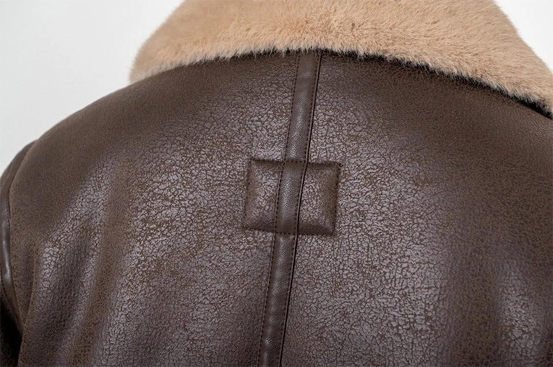 Puede incluir: Primer plano de una chaqueta de cuero marr&oacute;n con cuello de piel sint&eacute;tica beige. La chaqueta tiene una superficie texturizada y un detalle de parche cuadrado en la espalda. La costura es visible.