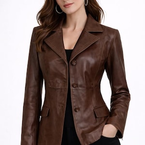 Puede incluir: Chaqueta de cuero marrón con un diseño clásico. La chaqueta presenta un cuello con solapas, cierre de botones y dos bolsillos delanteros. La chaqueta se lleva sobre un top negro y pantalones negros.