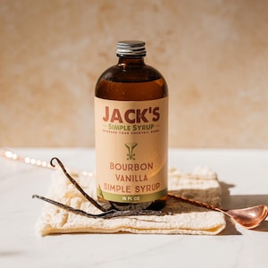 Puede incluir: Una botella de vidrio marrón de Jarabe Simple de Vainilla Bourbon de Jack's con tapa plateada. La etiqueta es de color beige con texto verde y marrón. Dos vainas de vainilla y una cuchara de cobre descansan sobre un paño color crema.