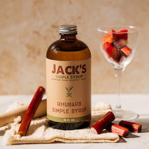 Puede incluir: Una botella de Jarabe Simple de Ruibarbo de Jack's, con una etiqueta. La imagen incluye una copa de cóctel con trozos de ruibarbo, tallos de ruibarbo y un mortero de madera. La botella de jarabe es marrón con una tapa plateada.
