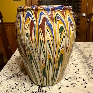 May include: A large, ceramic vase with a unique, swirled pattern. The vase features vertical streaks of blue, yellow, green, and maroon against a cream background. The rim has a wavy design. The vase is a decorative item.