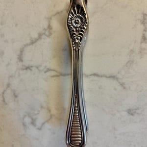 May include: A silver-toned metal sugar tong featuring an intricate embossed floral pattern at the top hinge and a series of horizontal ridges at the bottom gripping end. The utensil has a slender, elongated handle with a polished metallic finish.