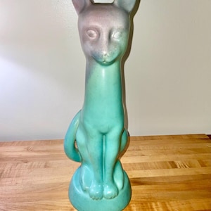 May include: A tall, stylized ceramic cat figurine featuring a smooth, matte finish. The sculpture is primarily a vibrant turquoise green, transitioning into a soft, muted purple and grey gradient across the ears and the top of the head.