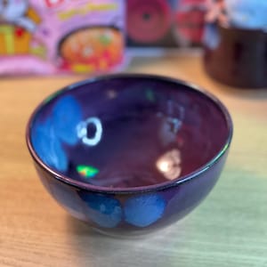 May include: A deep purple ceramic bowl with blue floral accents. The bowl has a glossy finish and a rounded shape. The interior reflects light, creating highlights. The bowl is sitting on a light-colored wooden surface.