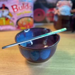 May include: A handmade ceramic bowl with a deep purple interior and blue accents, paired with a silver spoon. The bowl sits on a light-colored wooden surface, suggesting a kitchen or dining setting. A package of instant noodles is visible in the background.
