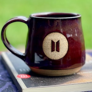 May include: A dark red ceramic mug with a glossy finish and a single handle. The front features a circular, textured beige medallion embossed with a dark brown vertical logo consisting of two parallel trapezoidal shapes.