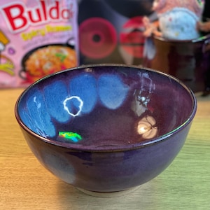 May include: A handmade ceramic bowl with a deep purple exterior and a vibrant blue interior. The bowl has a glossy finish and a rounded shape, perfect for serving soups or snacks. The bowl is sitting on a wooden surface.