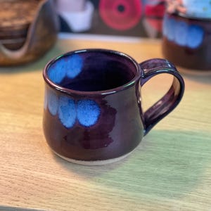 May include: A handmade ceramic mug with a deep purple glaze and blue accents. The mug has a rounded shape with a sturdy handle. The interior and the top rim of the mug are a deep purple color, with blue spots on the exterior. The mug sits on a light-colored surface.